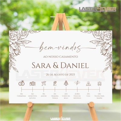 Placa de boas-vindas para casamento com decoração floral e texto em castanho num cavalete de madeira ao ar livre