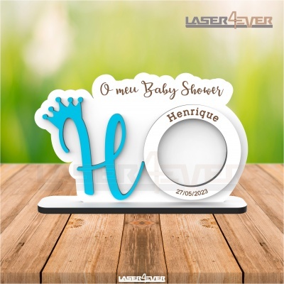 Peça decorativa para baby shower com letra azul e coroa, texto castanho e base preta sobre madeira