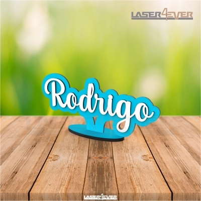 Peça decorativa azul com nome Rodrigo em branco sobre superfície de madeira com fundo verde e amarelo desfocado