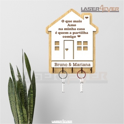 Suporte de chaves em madeira em forma de casa com texto em português e nomes Bruno & Mariana, duas chaves penduradas e planta verde ao lado