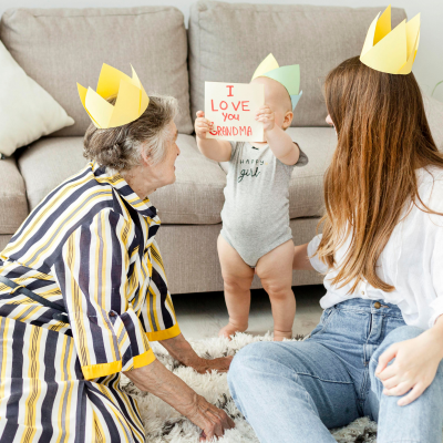 Criança com body cinzento segura cartão 'I LOVE YOU GRANDMA' entre duas adultas com coroas de papel amarelo sentadas no chão