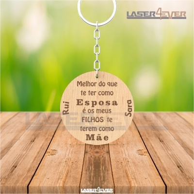 Chaveiro redondo em madeira clara com mensagem gravada em português e nomes Rui e Sara, pendurado sobre fundo de madeira e natureza verde.