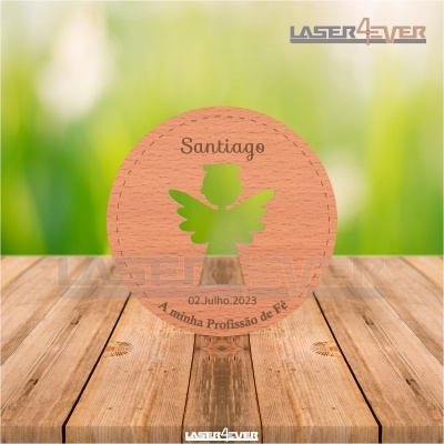 Peça redonda de madeira com recorte de anjo, texto gravado e fundo verde desfocado.