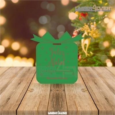 Placa decorativa verde em forma de presente com texto 'Feliz Natal' em vermelho, sobre madeira com fundo de árvore de Natal