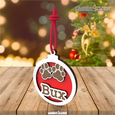 Pendente redondo vermelho com patas de animal e nome 'Bux', pendurado com cordão vermelho, árvore de Natal ao fundo