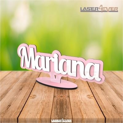 Decoração em acrílico com o nome Mariana em branco e rosa sobre base preta em mesa de madeira.