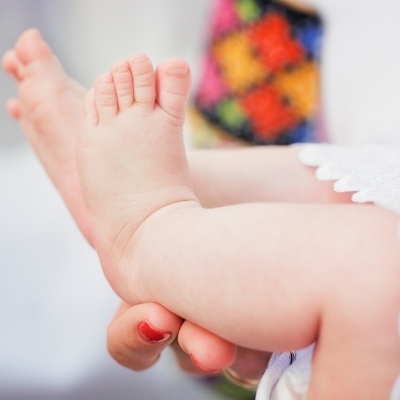 Pés e pernas de bebé segurados por mão com unhas vermelhas, fundo colorido desfocado