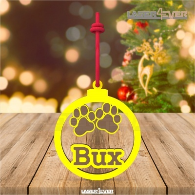 Enfeite amarelo com pegadas de patas e nome Bux pendurado numa mesa de madeira clara com decoração natalícia ao fundo