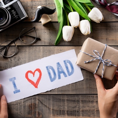 Papel com frase I coração DAD, tulipas brancas, presente embrulhado, óculos, câmara e objeto bigode numa mesa de madeira.