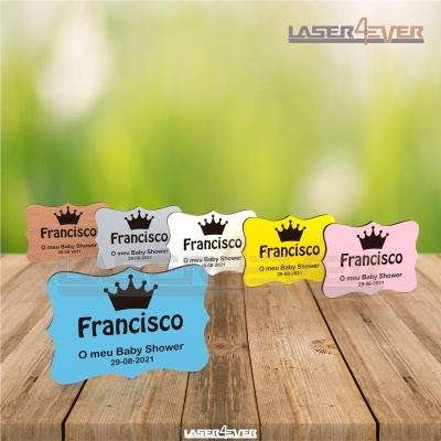 Etiquetas coloridas para baby shower com texto e coroa, sobre mesa de madeira clara com fundo verde desfocado