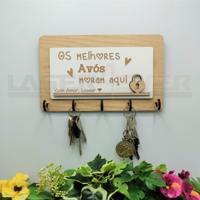 Suporte para chaves de madeira com placa branca gravada com texto sobre avós, pendurado numa parede branca com flores em baixo.