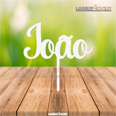 Topo de bolo em acrílico branco com a palavra 'João' em fundo desfocado verde e madeira.