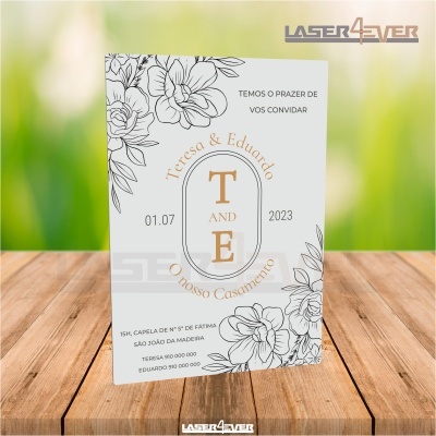 Convite de casamento com flores e texto em dourado e preto sobre fundo de madeira