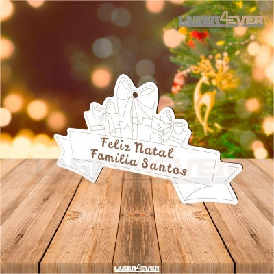 Placa decorativa branca em madeira com mensagem de Natal, sobre mesa de madeira com árvore de Natal ao fundo