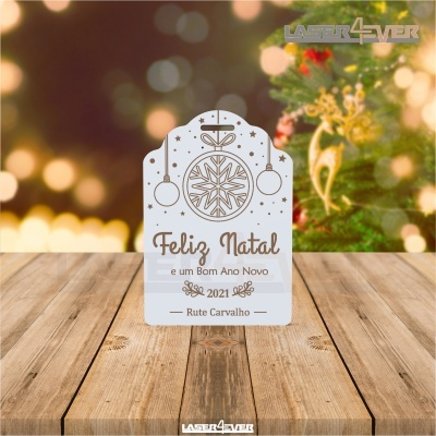 Placa decorativa de Natal branca com texto em castanho e enfeites, sobre superfície de madeira e árvore ao fundo