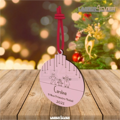Ornamento de Natal em madeira cor-de-rosa com fita vermelha e texto personalizado, sobre mesa de madeira e fundo natalício.