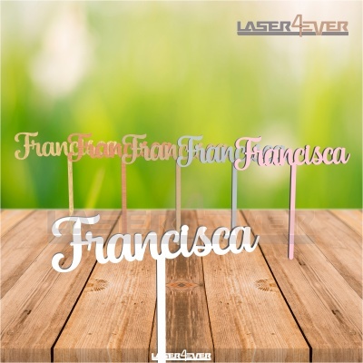 Topper decorativo com o nome Francisca em várias cores e materiais sobre mesa de madeira com fundo desfocado
