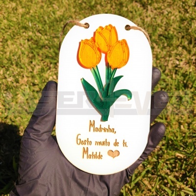 Placa oval branca com tulipas amarelas e texto em amarelo, segurada com luva preta sobre relvado.