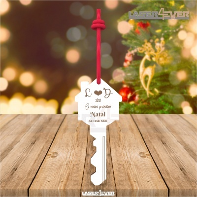 Chave pendurada em cordão vermelho com etiqueta de casa branca com texto de Natal sobre superfície de madeira e árvore de Natal ao fundo.