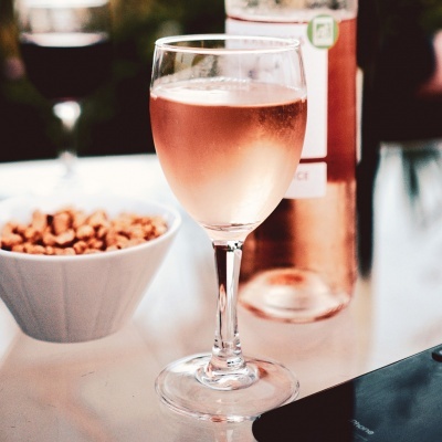 Taça com vinho rosé, garrafa e petiscos sobre mesa branca