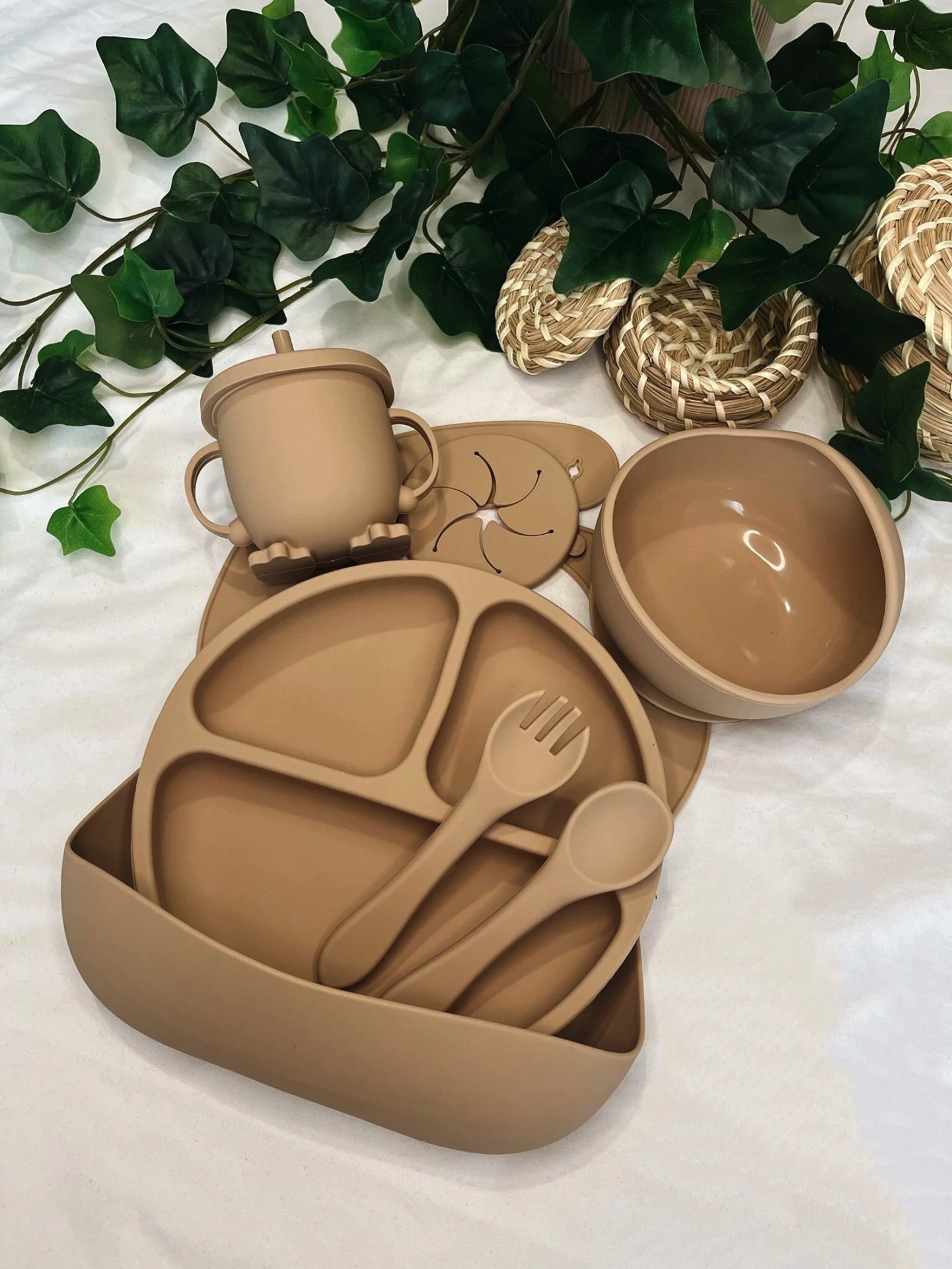 Conjunto de utensílios infantis em silicone bege com prato dividido, colher, garfo, tigela e copo, sobre tecido branco com folhas verdes