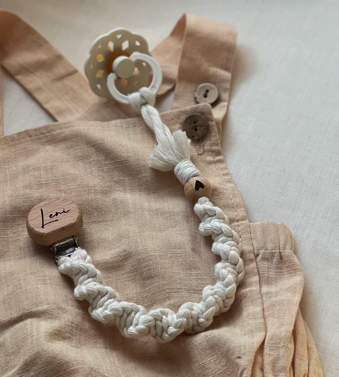 Prendedor de chupeta branco com contas de madeira sobre roupa bege com botões.