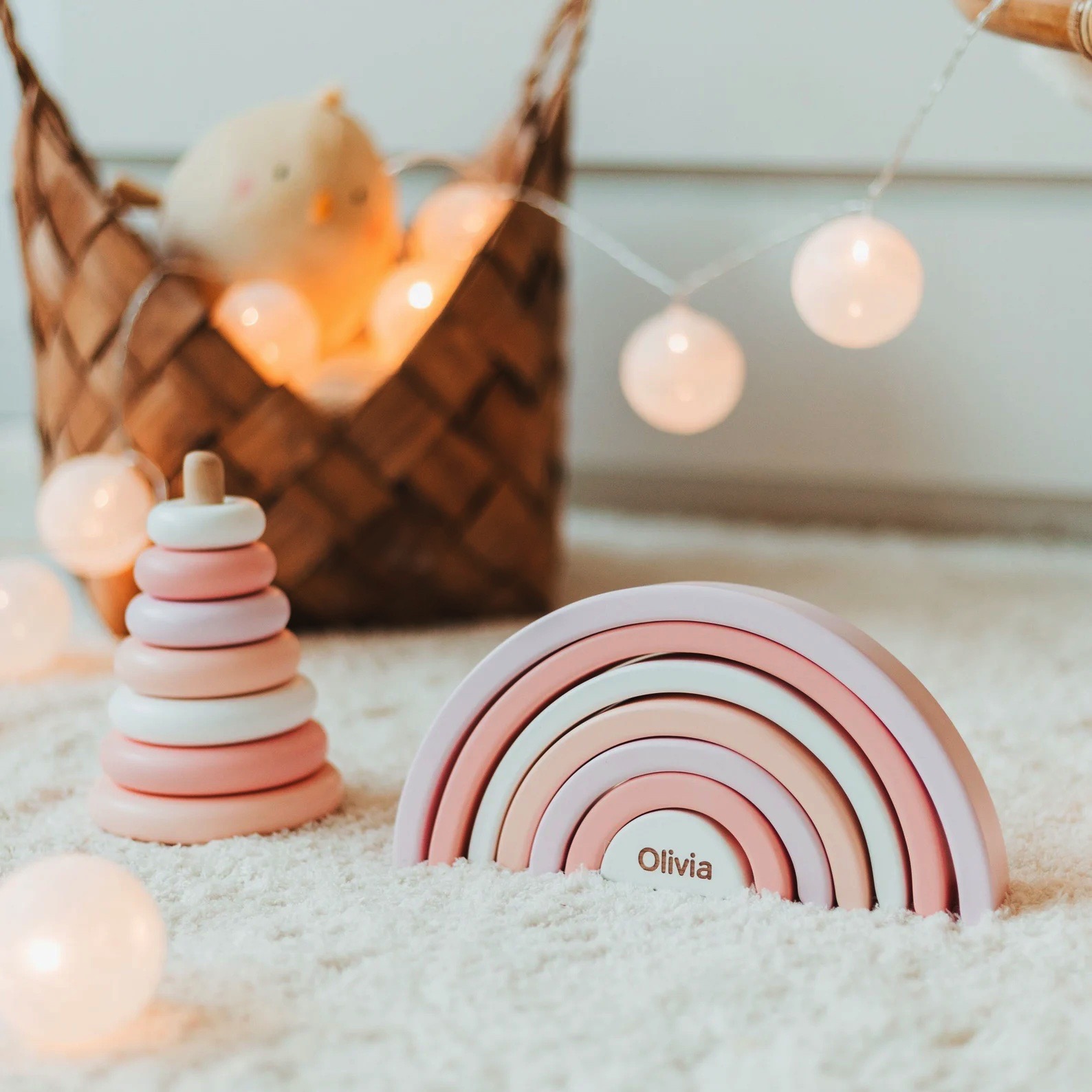 Brinquedos de madeira coloridos em tons rosa e bege sobre tapete branco com luzes decorativas e cesta ao fundo.