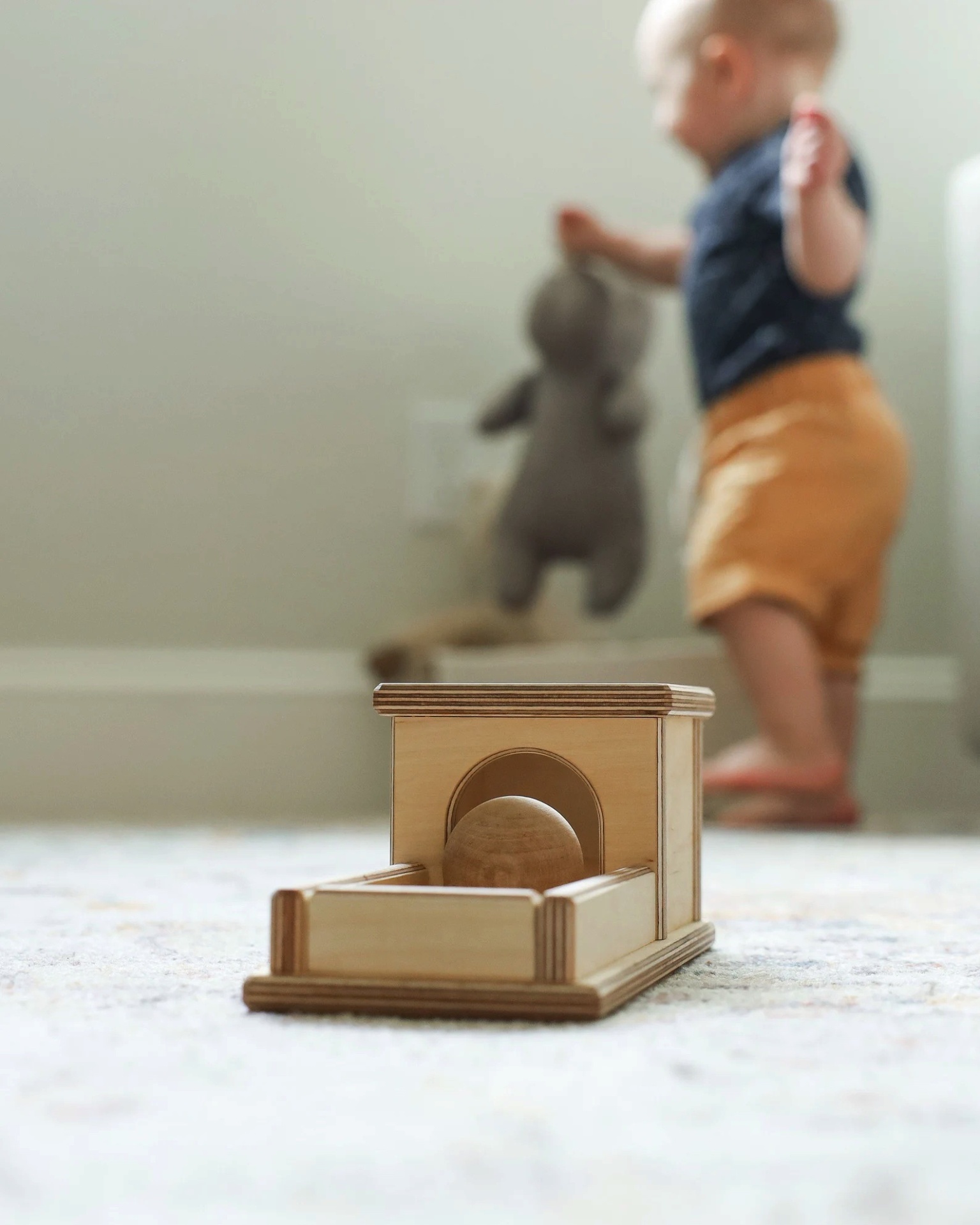 Brinquedo de madeira com túnel e bola, criança ao fundo com peluche cinzento