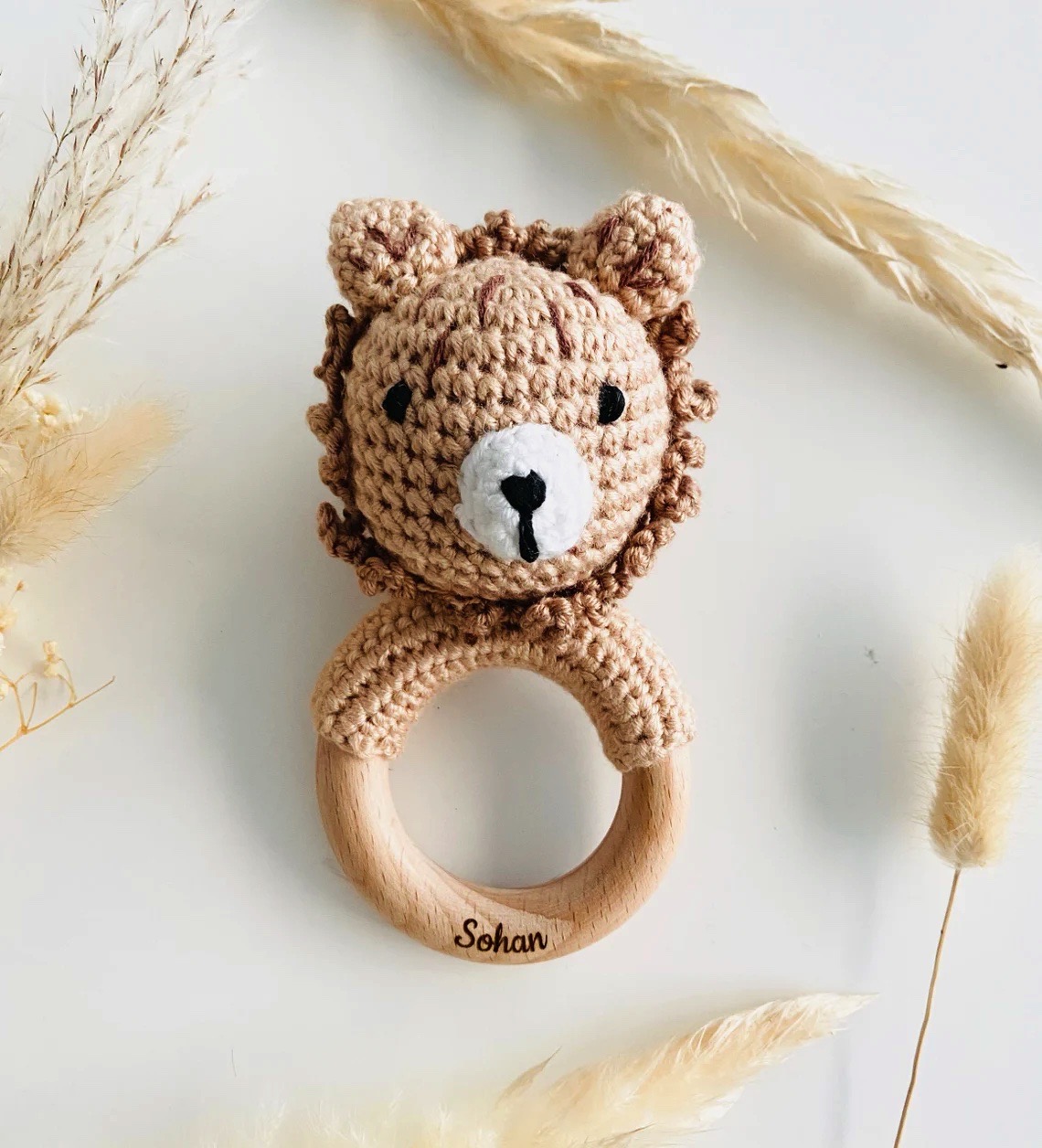 Chocalho de bebé com argola de madeira e cabeça de leão em crochet num fundo branco com ramos secos
