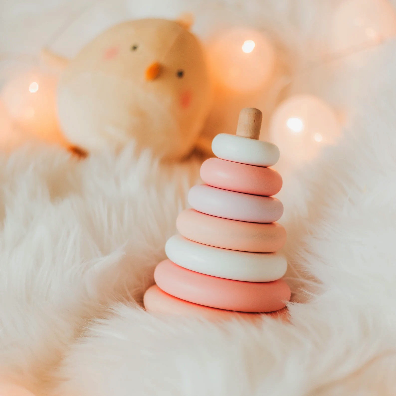 Brinquedo empilhável de madeira em tons rosa e branco sobre tecido felpudo branco com peluche de pintainho ao fundo