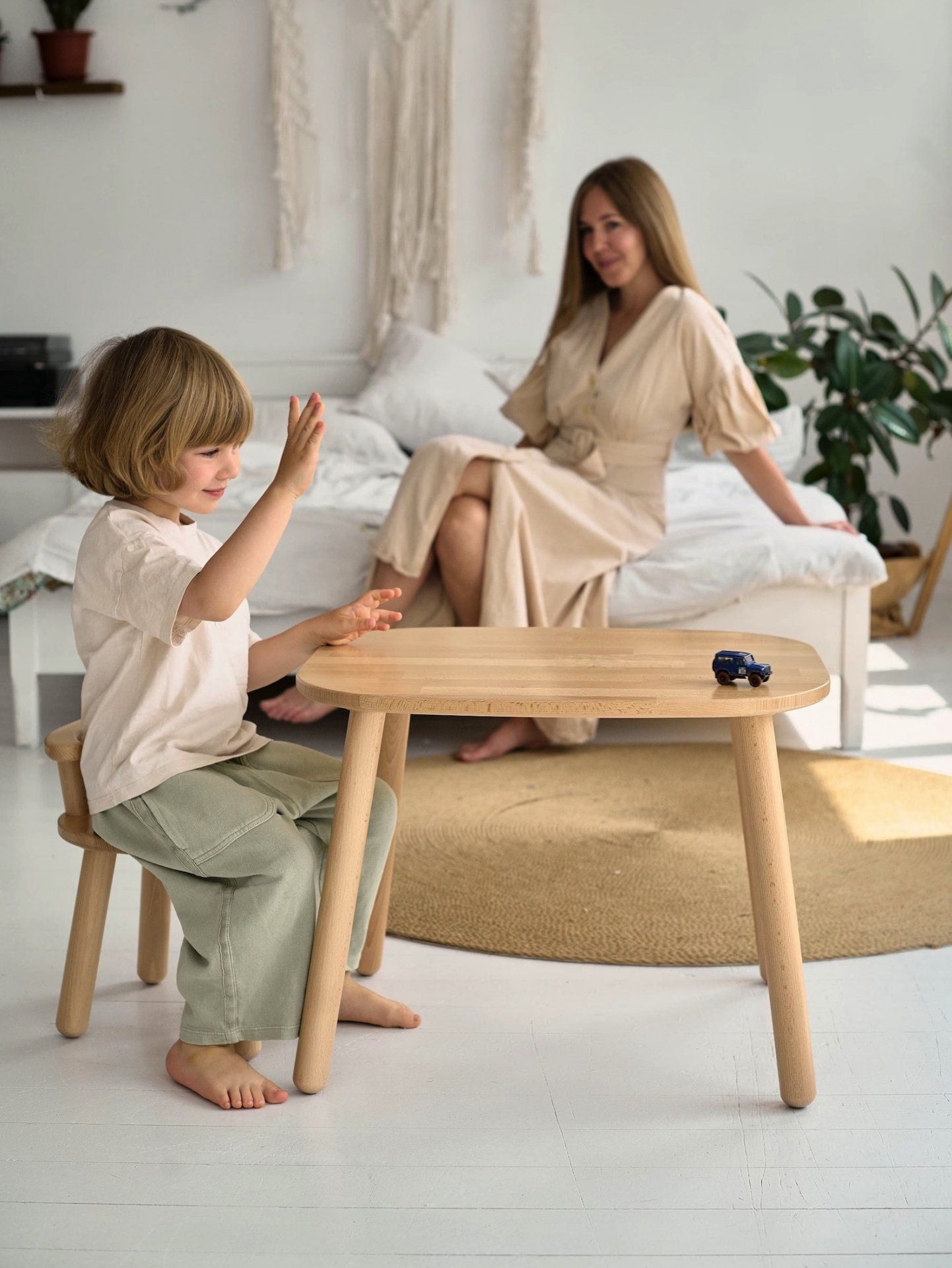Criança sentada numa pequena cadeira de madeira clara à frente de mesa baixa oval de madeira com mulher sentada ao fundo em sofá branco