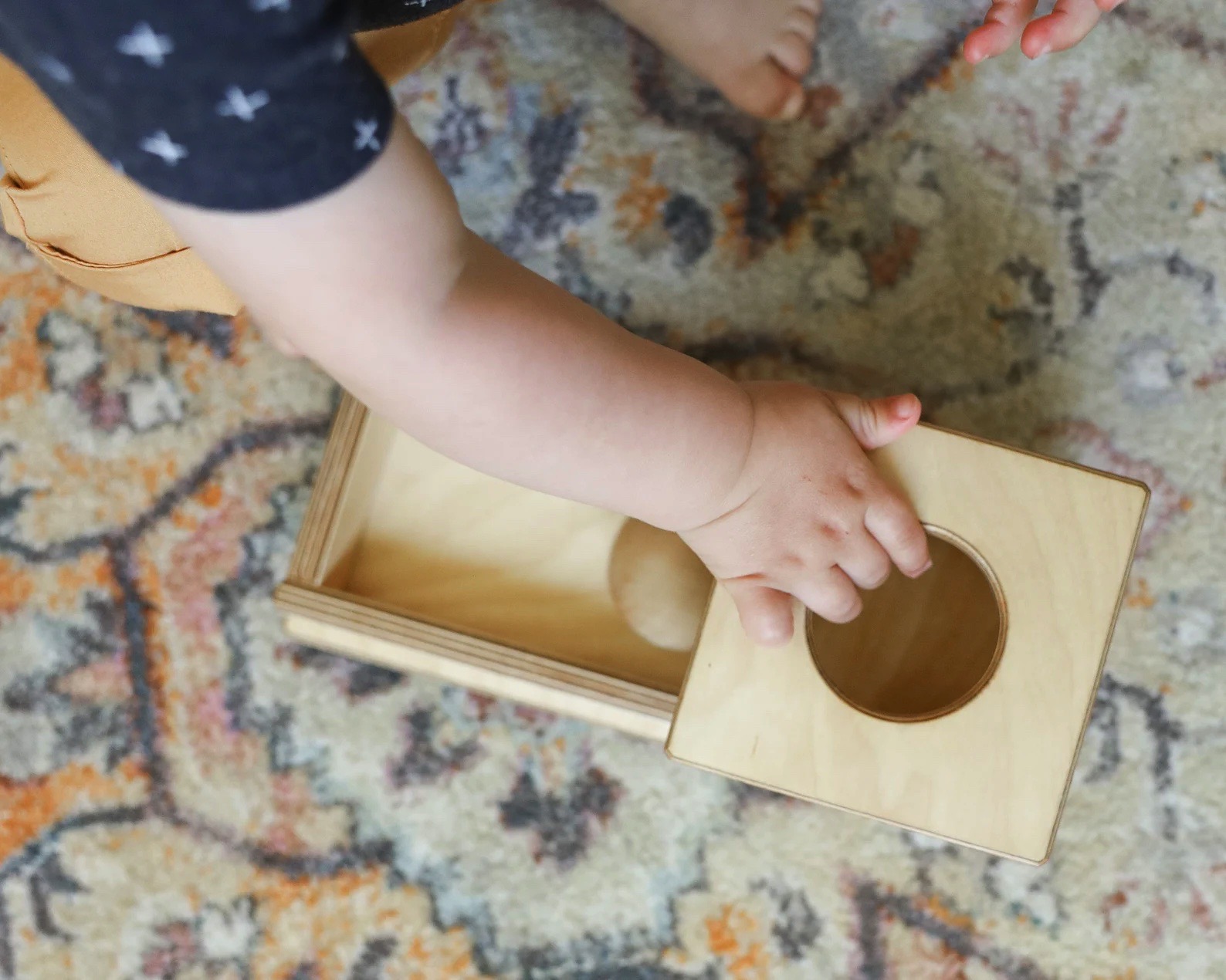 mão de criança a pegar tampa de madeira numa caixa sobre tapete colorido