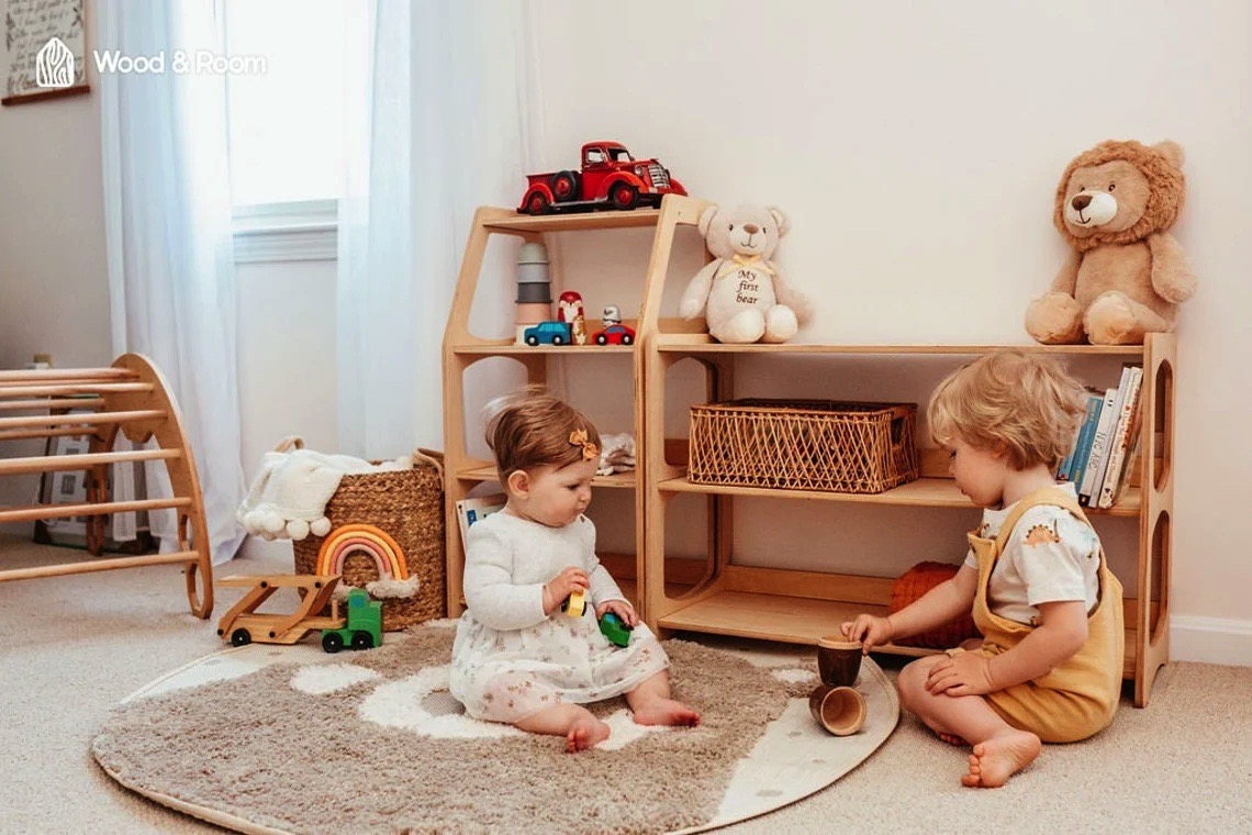 Dois bebés brincam num tapete cinzento com móveis e brinquedos de madeira clara numa sala iluminada.