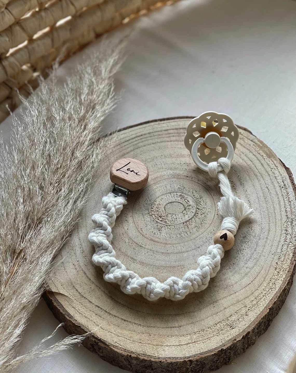 Chupeta branca com fita trançada e contas de madeira numa base de madeira circular.