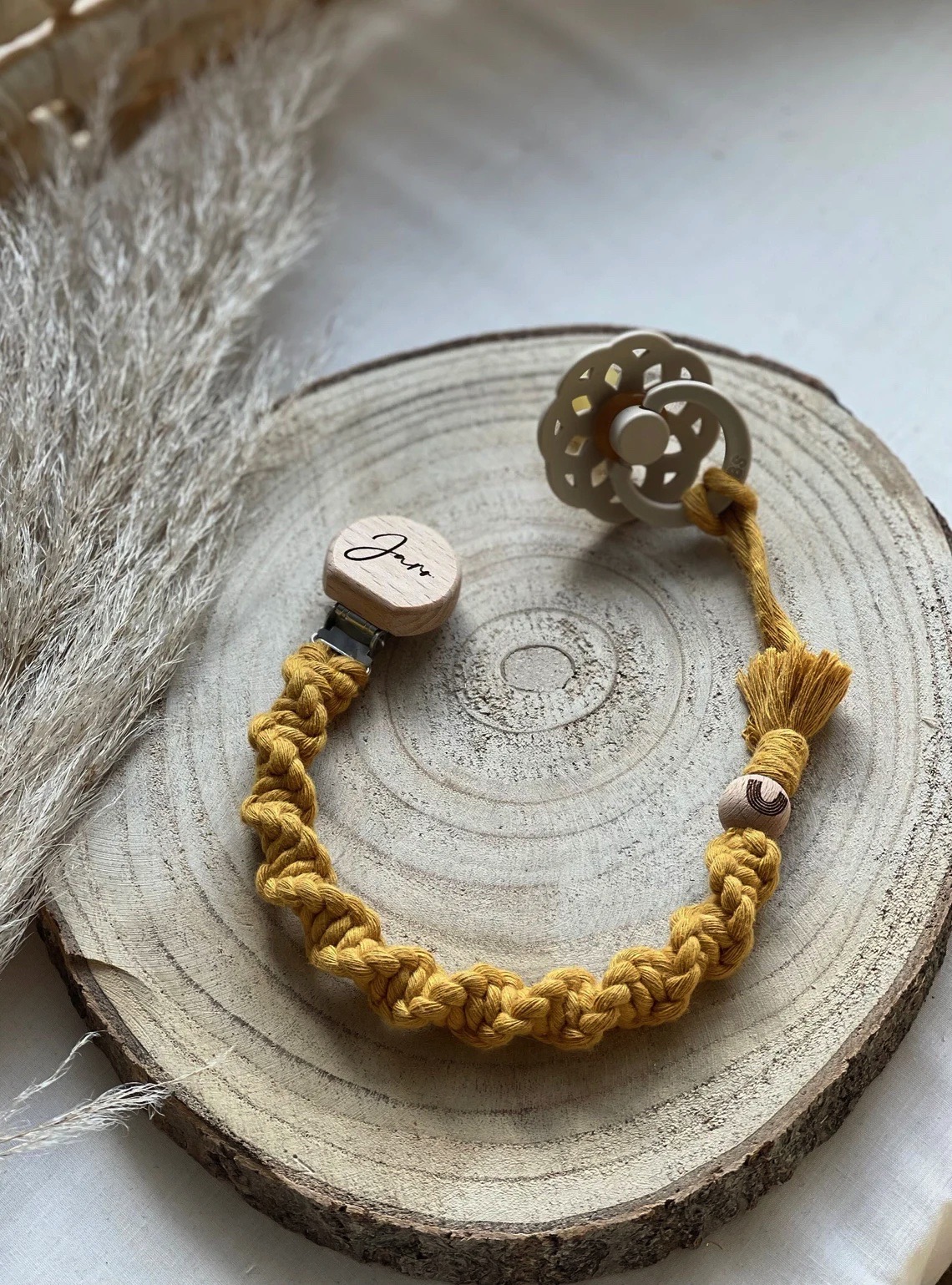 Prendedor de chupeta amarelo em fio trançado com clip de madeira e chupeta bege em forma de flor sobre base de madeira redonda