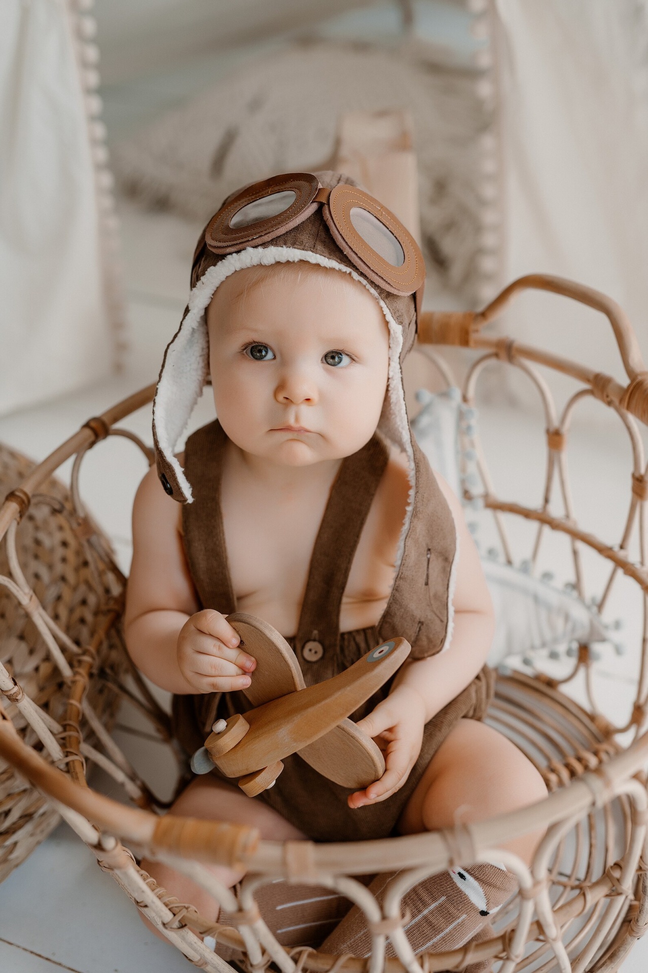 Bebé com chapéu de aviador e macacão castanho sentado numa cadeira de rattan segurando um brinquedo de avião de madeira.