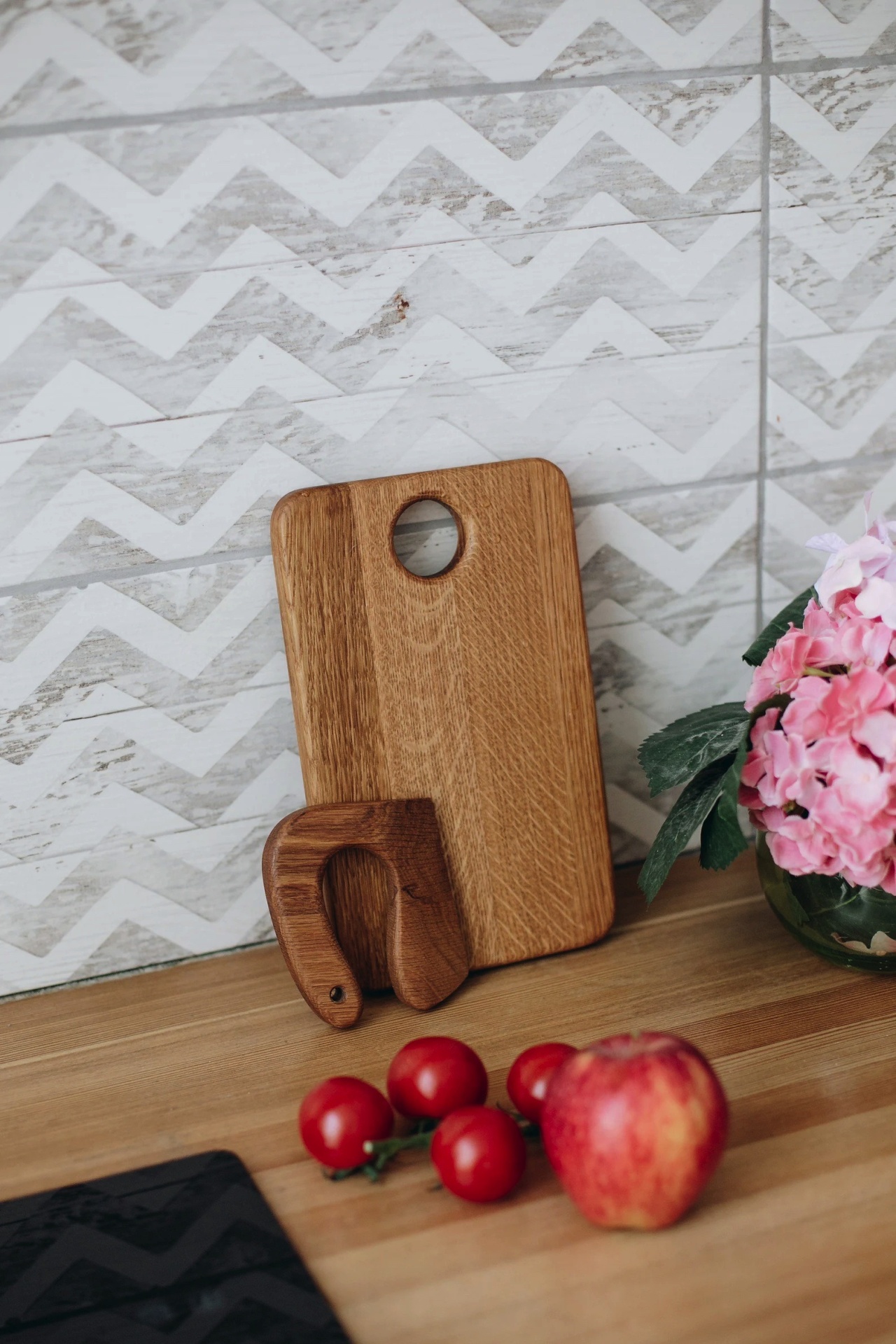 Tabua de cortar em madeira com orificio, suporte em madeira, tomates cherry, maçã e flores rosa num vaso