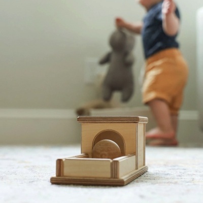 Brinquedo de madeira com túnel e bola, criança ao fundo com peluche cinzento