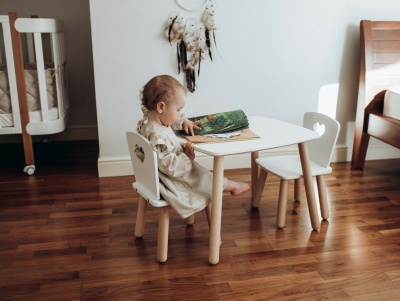Criança sentada numa cadeira branca a olhar um livro verde numa mesa branca com pernas de madeira