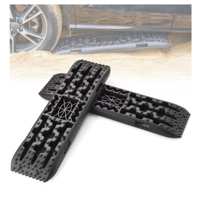 Placas de tracção negras com textura ondulada sobre chão arenoso perto de roda de carro preta