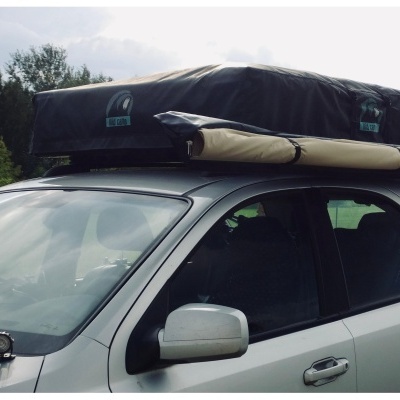 SUV cinzento claro com tenda de teto preta e toldo bege ao lado, em ambiente ajardinado