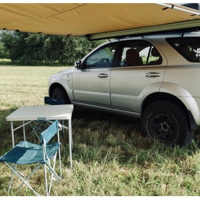 SUV cinza com toldo amarelo, mesa branca dobrável e cadeira azul em área gramada