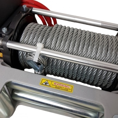 Rolo metálico de cabo de aço prateado numa estrutura cinzenta com rótulo de aviso.