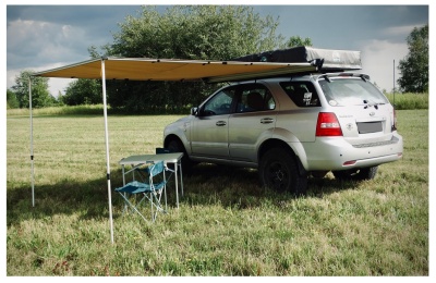 Carro 4x4 prata com toldo amarelo e mesa com cadeiras azuis em campo