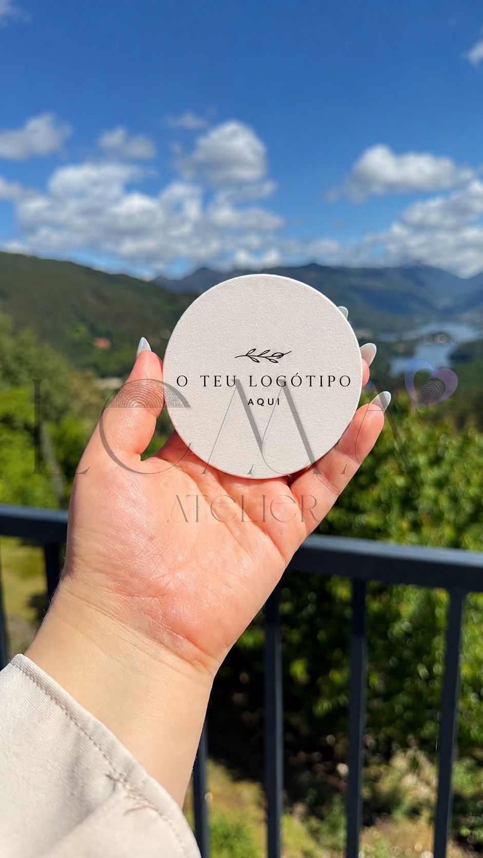 Base para Copos — Personaliza tu! Objeto redondo branco com texto O TEU LOGÓTIPO AQUI segurado por mão contra céu azul e vegetação