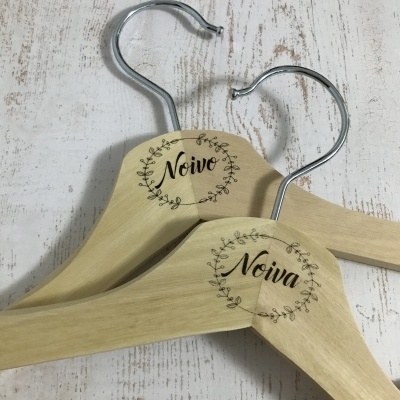 Dois cabides de madeira clara, um com 'Noivo' e outro com 'Noiva', sobre fundo de madeira branca desgastada