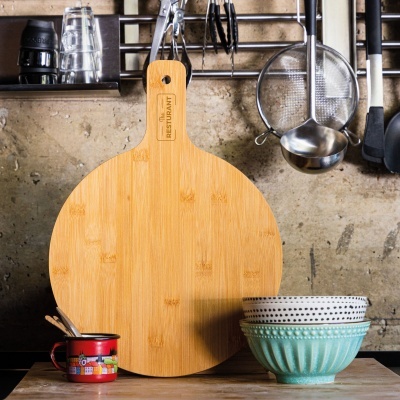 Tabuleiro redondo de madeira clara com cabo e inscrições, utensílios de cozinha pendurados e tigelas cerâmicas coloridas numa bancada