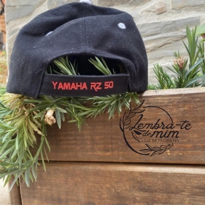 Boné preto com texto vermelho Yamaha RZ 50 colocado em banco de madeira com plantas verdes e logotipo Lembra-te de mim