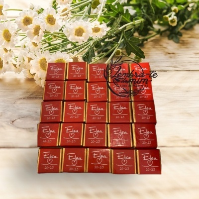 Pequenos chocolates vermelhos com texto Elsa 20-23 em superfície de madeira com flores brancas ao fundo