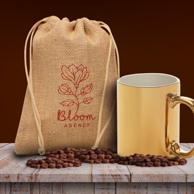 Saco de estopa com estampa vermelha e caneca dourada sobre mesa de madeira com grãos de café